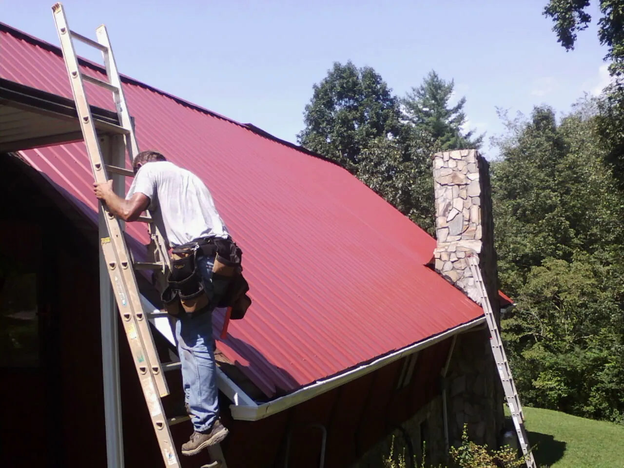 Roofer on a ladder beside a red metal roof and stone chimney — Soffit & Fascia Repair by Roofing Brothers in Quinault